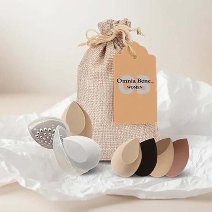 A set of teardrop makeup sponges with clear plastic, breathable protective cases, and a beige burlap drawstring pouch with a tan tag reading 'Omnia Bene Women sitting on crinkled white tissue paper