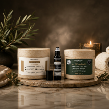 Three wellness products from Omnia Bene arranged on a round stone tray: a large beige body butter jar, a small black roller bottle, and a larger beige foot soak jar with a dark green label. The scene is styled with olive branches, a lit candle, and a folded towel against a warm brown background.