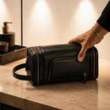 A man's hand resting on an Omnia Bene Puccian Nappa leather dopp kit on a marble bathroom counter.